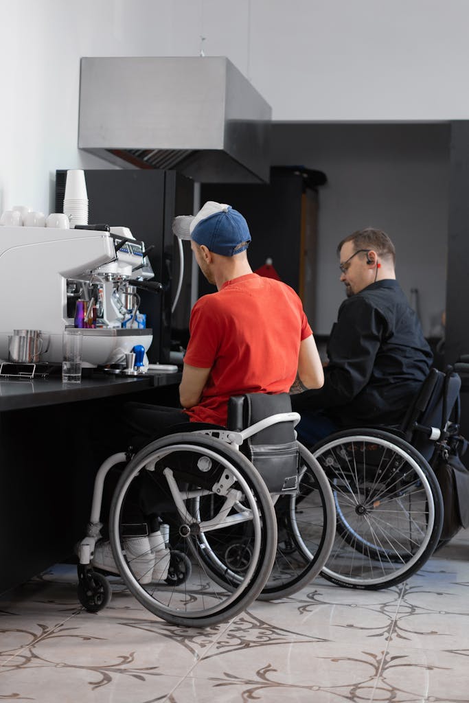 Two men sitting in wheelchairs at a coffee shop counter, engaging with a barista in an indoor setting.